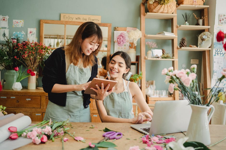 Two women in a floral workshop or shop, one standing and the other seated at a wooden table adorned with pink and red flowers, wrapping paper, and floral tools. The standing woman, with shoulder-lengt