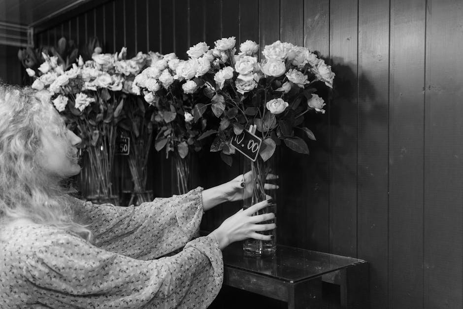 A woman with curly blonde hair, wearing a patterned long-sleeve blouse, carefully arranges a large bouquet of fresh white roses in a tall clear glass vase. The bouquet features numerous blooms with fu