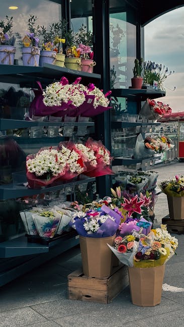 A display of various floral bouquets arranged on metal shelves outside a flower shop, including arrangements of white daisies, pink lilies, and mixed colorful blooms wrapped in purple, pink, and brown