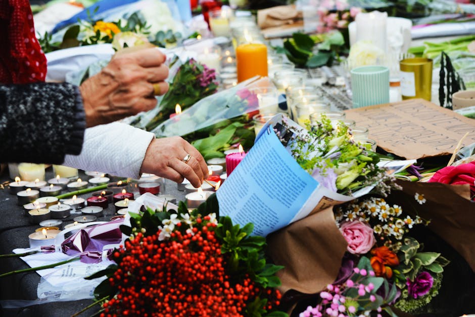 Close-up of a floral tribute scene with hands arranging flowers on a table. The arrangement includes a bouquet of pink roses, white daisies with yellow centers, and purple flowers wrapped in brown pap