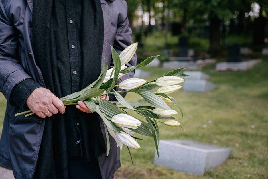 A person dressed in a dark coat and shirt holding a bouquet of fresh white lilies with green leaves, arranged with the stems bundled together. The bouquet is positioned in front of the person's torso,