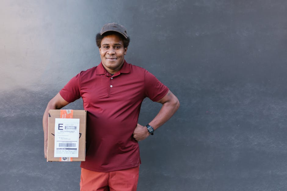 A male flower delivery courier with medium brown skin and curly dark hair, wearing a maroon polo shirt, matching cap, and red trousers, standing outdoors against a dark grey wall. He is smiling and ho