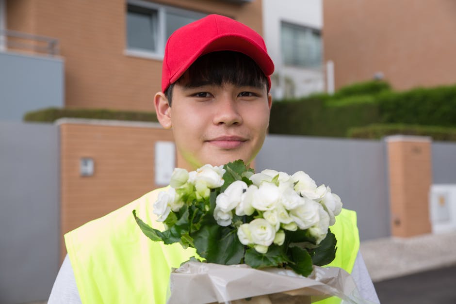 A young male delivery person wearing a red cap and a high-visibility yellow vest is standing outdoors in front of modern residential buildings with grassy landscaping. He is holding a bouquet of fresh